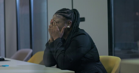 African American businesswoman sits with face buried in hands at office desk, struggling with...