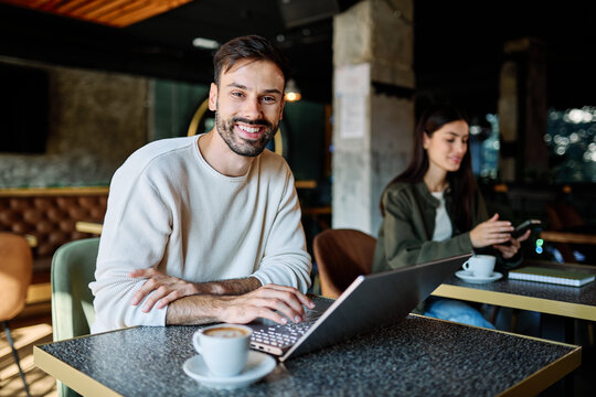 Man and woman smiling, holding coffee while working on a laptop and using a mobile phone  in a cafe