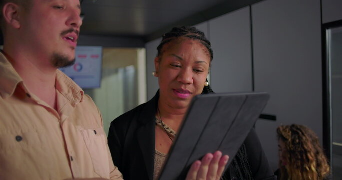 African American businesswoman smiling at digital tablet with young male colleague, collaborative moment in modern office during team meeting