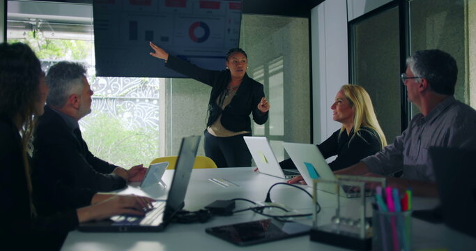 African American businesswoman pointing to data chart on screen while delivering presentation to engaged colleagues in modern glass walled office