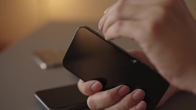 Person hands holding smartphone with cracked screen while using ejector tool to access sim card tray, close-up. Concept of telecom, repair, device management. Shooting in slow motion.