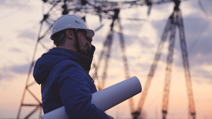 Obraz premium An electrician on the background of high towers of power plants looks at the project for the development of an electrical structure, the expansion of the electrical voltage of volts in the wires.