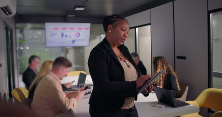 African American businesswoman standing in office using digital tablet, focused on work while colleagues collaborate at conference table in background