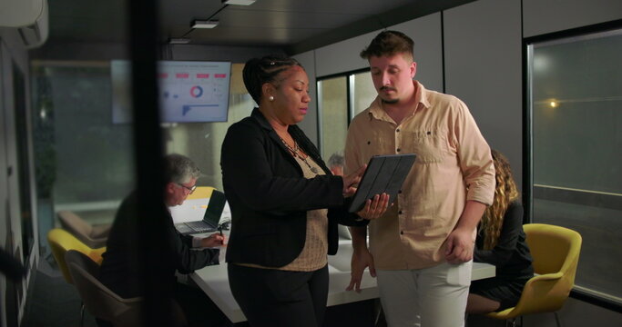 African American businesswoman showing tablet to young male colleague during office meeting, teamwork and analysis in front of large data display