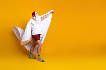 Santa in pajama with red hat and long beard pulls a flowing white cape against a bold yellow background in a playful holiday scene