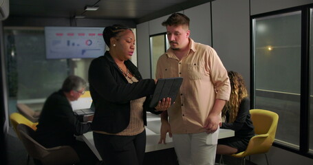 African American businesswoman showing tablet to young male colleague during office meeting, teamwork and analysis in front of large data display