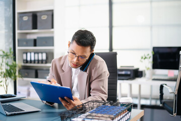 Asian businessman talking on smartphone while working on laptop at modern office desk, symbolizing business communication and corporate success.