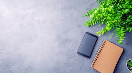 A modern workspace setup featuring a smartphone, a spiral-bound notebook, and a lush green plant on a textured gray background.