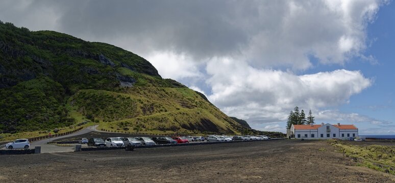 Car park with many cars next to a white house and thermal bath in a lava landscape under a cloudy sky, Termas da Ferraria, Sao Miguel Island, Azores, Portugal