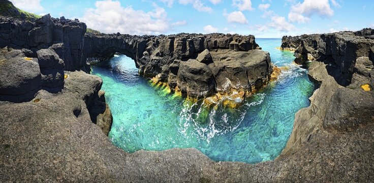Panoramic view of rugged lava rock formations with a natural arch over turquoise sea under blue sky, A Porta do Diabo, Sao Miguel Island, Azores, Portugal
