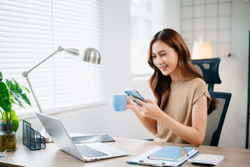 Smiling Asian businesswoman holding smartphone and coffee cup in bright office. Perfect for business lifestyle, digital work, communication