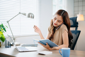 Asian businesswoman multitasking with phone and notebook in bright office. Perfect for business, communication, consulting, productivity, and professional work themes.