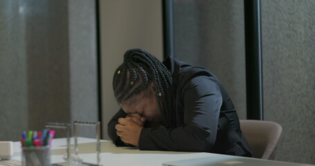 African American corporate employee leans forward at table with face hidden behind hands, conveying...