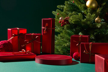 Red presents and Christmas ornaments are arranged on a green table with a tree behind them. The...