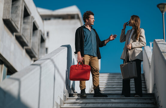 Two young professionals converse on a sunny outdoor stairway, each carrying a bag. The scene conveys business, style, and casual professionalism in an urban setting.