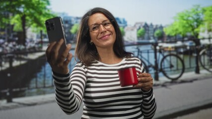 Brunette woman taking selfie with smartphone while holding bright red mug on urban street canal walkway with railing and bicycle in view; playful. - Powered by Adobe