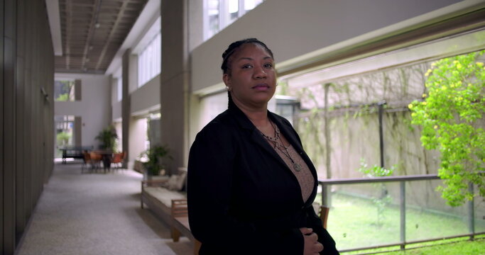 African American professional woman standing confidently in modern office hallway, looking at camera with calm expression beside large glass windows and greenery outside - Powered by Adobe