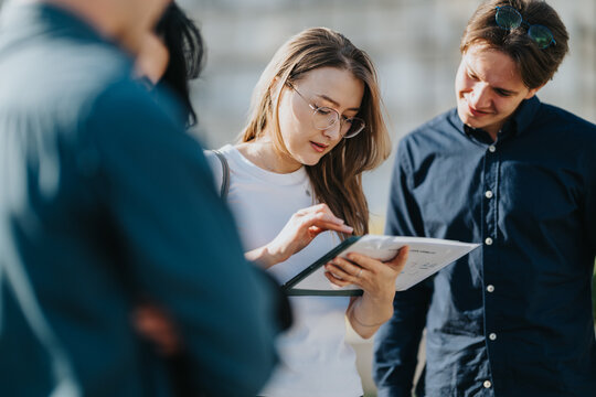 A group of young professionals stands outside, examining a tablet and a clipboard. They discuss ideas, share insights, and collaborate in a sunny, casual business moment.