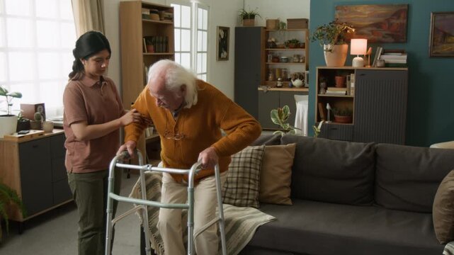 Slow motion shot of Latin American young woman assisting senior man trying to get up from couch at home using walker with shaking hands, copy space