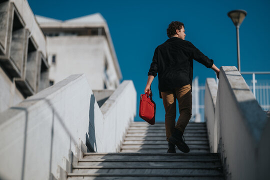 A man in casual business attire climbs a concrete stairway outdoors, carrying a red bag. The urban setting features modern architecture and a bold blue sky, conveying travel, work, and momentum. - Powered by Adobe
