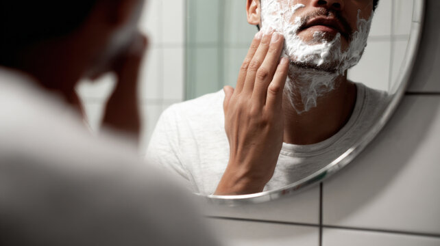 A person looking into the mirror with shaving cream on their face and applying it 