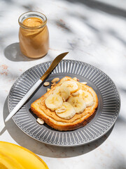 Peanut butter toast and banana on a dark plate on a marble background with fruits