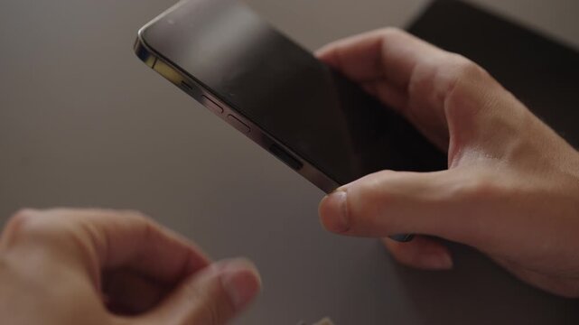 Close-up of male hands holding and inserting nano sim card into new smartphone tray sitting on desk, illustrating mobile service transfer, setup, and device upgrade connectivity, slow motion.