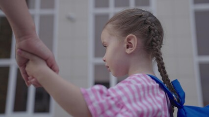 little girl with backpack holds her mother by the hand and goes to school, kid with mom go to school in the school yard, child with school bag hurries to first lesson, preparatory preschool education.