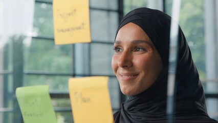 Satisfied islamic business woman analyzing stickers on glass in office Muslim businesswoman in hijab company CEO entrepreneur female girl reading sticky notes data plans smiling management work task