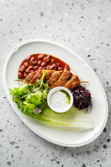 ground meat kofta kebabs on a skewer, served with a side of kidney beans in tomato sauce, mixed green salad, red cabbage, and a creamy yogurt sauce dip on a white plate