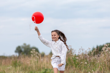 Girl runs joyfully in a field while holding a red balloon on a sunny day