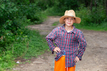 Young boy in a straw hat stands proudly on a forest path surrounded by greenery during daytime