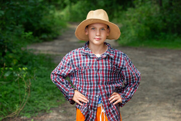 Young boy in a straw hat wearing plaid shirt stands confidently on a forest path during sunny afternoon