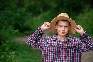 A young boy stands confidently in a plaid shirt and straw hat. He is smiling while posing in a lush green environment, enjoying a sunny day outdoors.