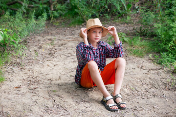 A young boy sits on the ground in a forest, dressed in a plaid shirt and bright orange shorts. He wears a straw hat and looks contemplative, surrounded by nature.