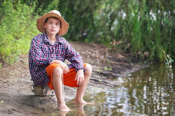 Young boy wearing a straw hat sitting by the riverbank during a sunny day while enjoying nature