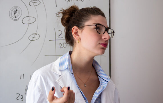 Female teacher wearing a lab coat and glasses, explaining complex chemistry concepts on a whiteboard during a science class, demonstrating education and learning