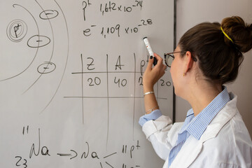 Female chemistry teacher explaining atomic structure and chemical equations on a white board during a science lesson, educating students in a classroom setting