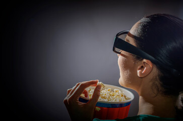 A woman wearing 3D glasses is viewed from the side, illuminated by the light of a screen as she eats popcorn from a striped bucket in a dark theater or home cinema setting.