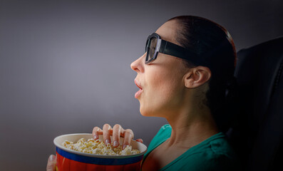 A woman wearing 3D glasses is viewed from the side, illuminated by the light of a screen as she eats popcorn from a striped bucket in a dark theater or home cinema setting.