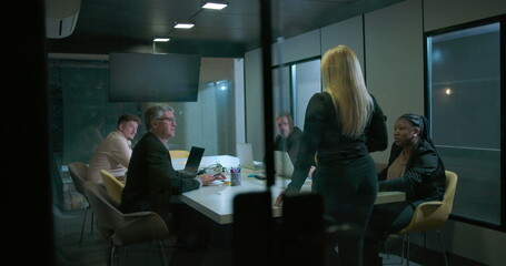 Back view of woman entering business meeting as team members look up in modern glass conference room during evening work session