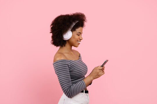 Smiling African American woman wearing headphones and using smartphone on pink background. Concept of music, technology, and relaxation in daily lifestyle - Powered by Adobe