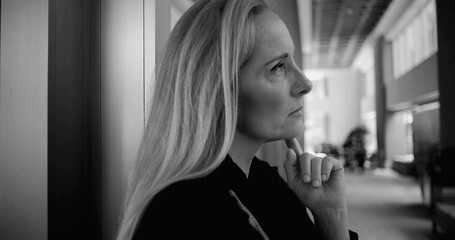 Black and white image of woman looking upward in contemplation while standing against wall in spacious corridor with office furniture in soft focus