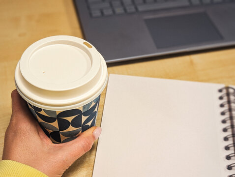 hand holding a geometric coffee cup next to a blank notebook and laptop on a wooden desk