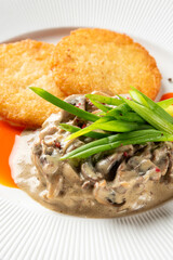two golden-brown potato pancakes served on a white plate with a creamy mushroom sauce and green onion garnish on a speckled grey surface.