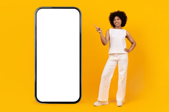 Smiling young African American woman standing next to big blank smartphone mockup on yellow background. Modern digital technology and app concept