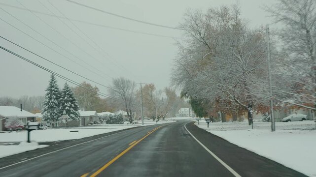 Snowy American suburban road with winter trees, yellow autumn foliage and houses on both sides, viewed through a moving car while driving forward.