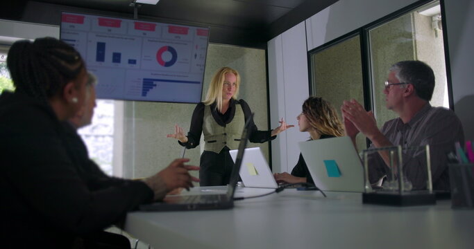 Blonde businesswoman giving presentation in front of data screen as coworkers watch and interact in professional modern office setting