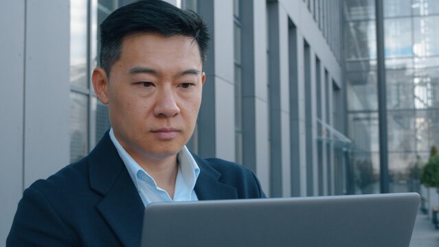 Serious Asian middle-aged business man with laptop in city working focused Korean businessman Chinese Japanese entrepreneur employer financial analyst with computer typing remote job technology using