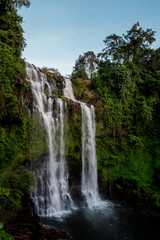 Obraz premium A powerful multi-tier waterfall cascades down rocky cliffs surrounded by dense tropical greenery. The flowing white water creates dynamic motion in a dramatic, scenic outdoor landscape.